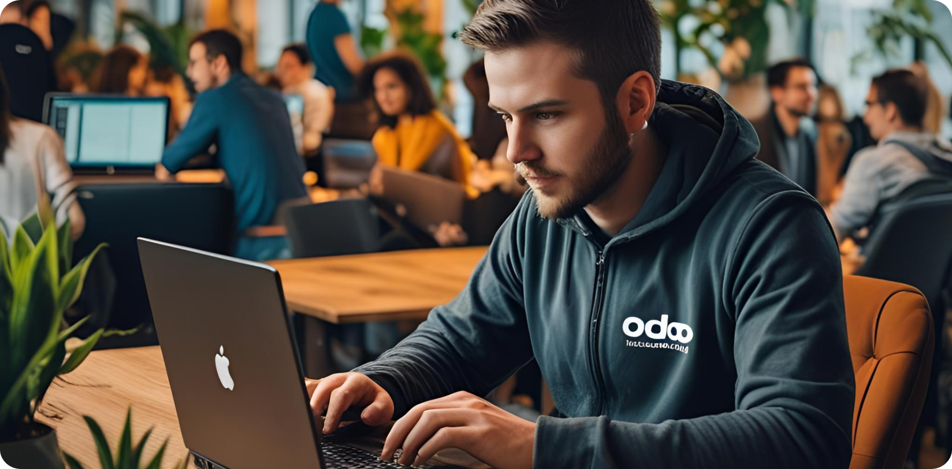 A focused young man in a dark hoodie with the Odoo logo works on a MacBook at a modern co-working space. Around him, groups of people collaborate and work on laptops in a well-lit, plant-filled environment, creating a vibrant and productive atmosphere.