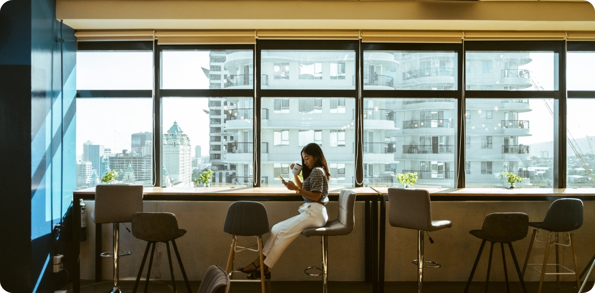 A young professional enjoying coffee while working on her phone in a bright, modern co-working space with city views, surrounded by stylish bar stools and large windows. Perfect for co-working space Makati or co-working space Cebu, this flexible workspace offers a productive and inspiring environment for freelancers, entrepreneurs, and teams.