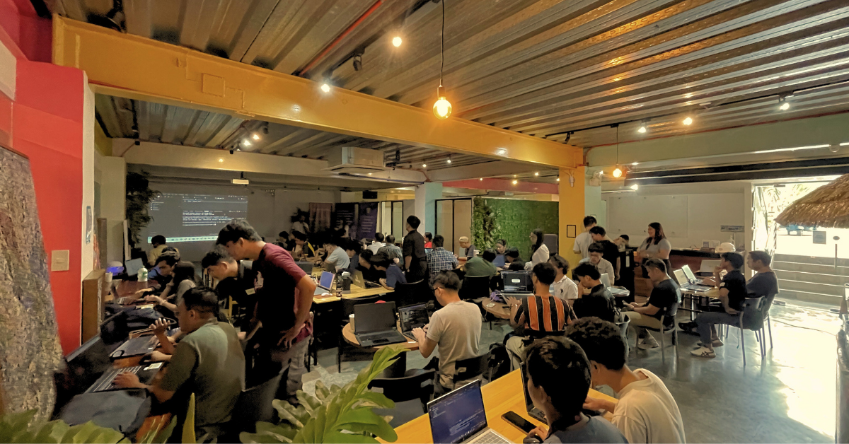 A wide shot of the "Build with AI" workshop at The Company Cebu, showing participants seated at laptops, deeply engaged in hands-on AI development. The room is bright and collaborative, with cables and equipment spread across tables. A facilitator presents at the front, introducing advanced agentic AI concepts. The scene reflects a transformative atmosphere—participants are building generative agents and multi-agent systems with Google's Agent Development Kit, bridging local insight and global technologies.