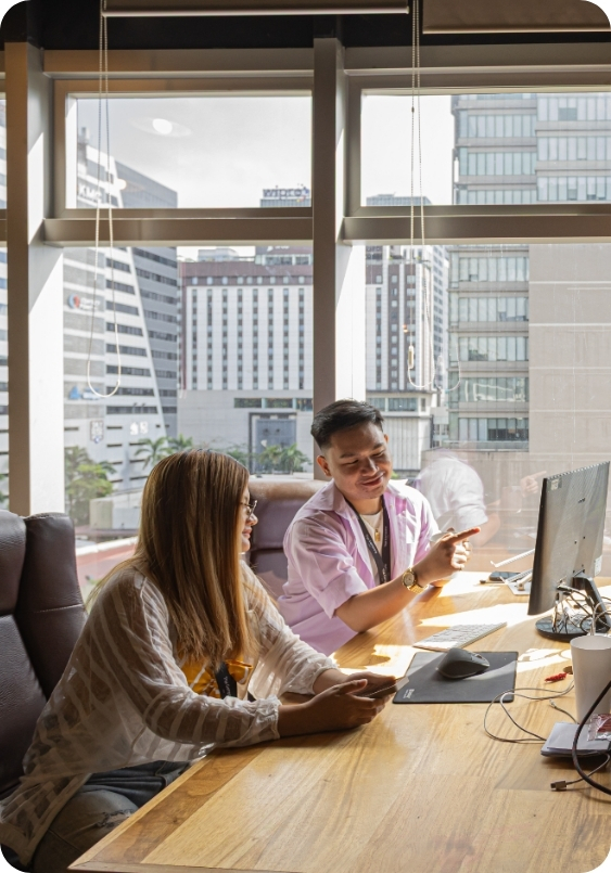 A man and woman collaborate at a shared desk, reviewing a computer screen with floor-to-ceiling windows overlooking Makati’s business district. The Company Makati’s Virtual Office solution makes professional teamwork in Makati effortless and inspiring.