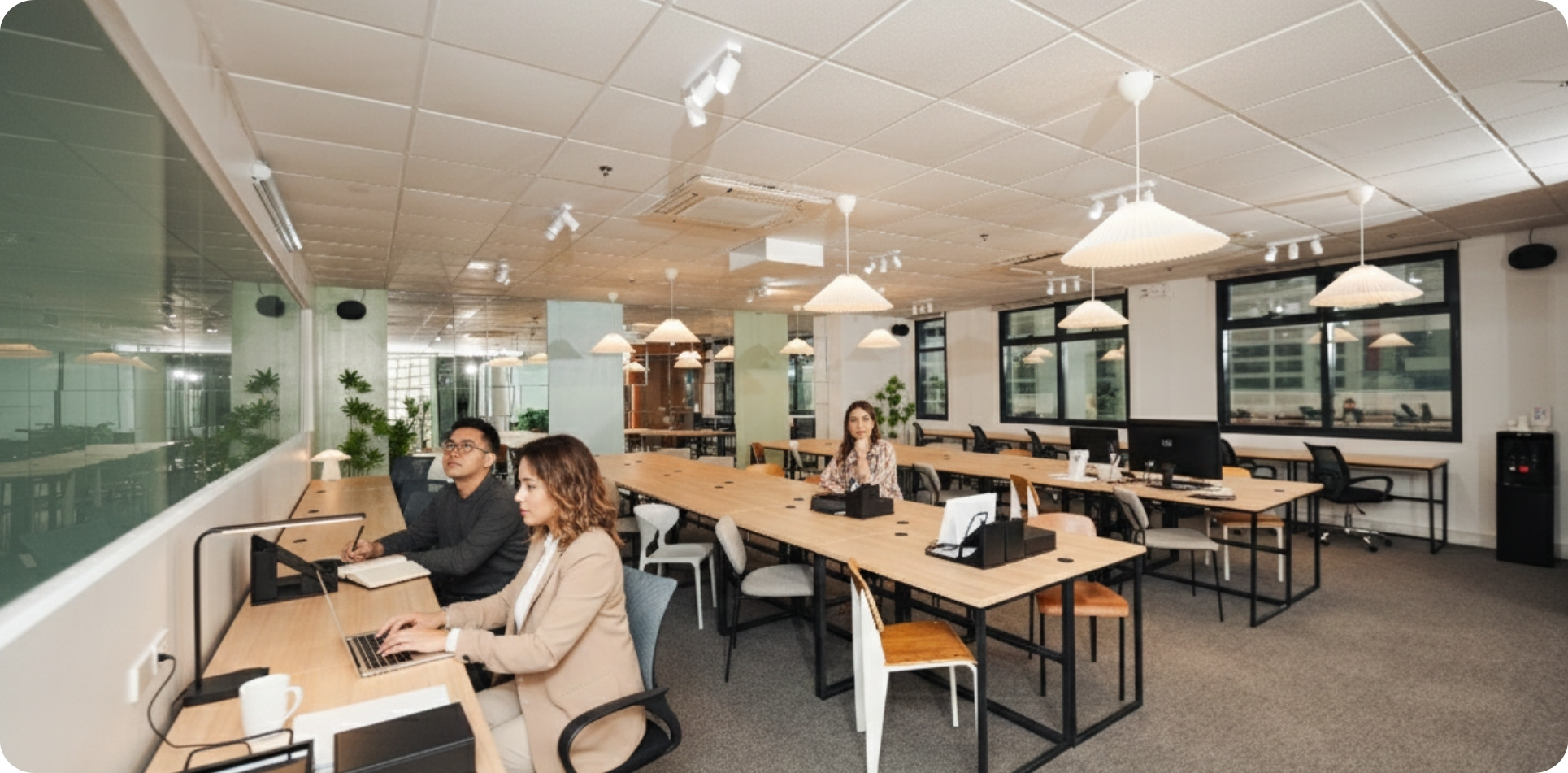 A bright, modern, and spacious plug and play office in Makati with several individuals working under EOR comfortably at long wooden desks. The office features contemporary pendant lighting, large windows, and some green plants, creating a productive and inviting atmosphere.